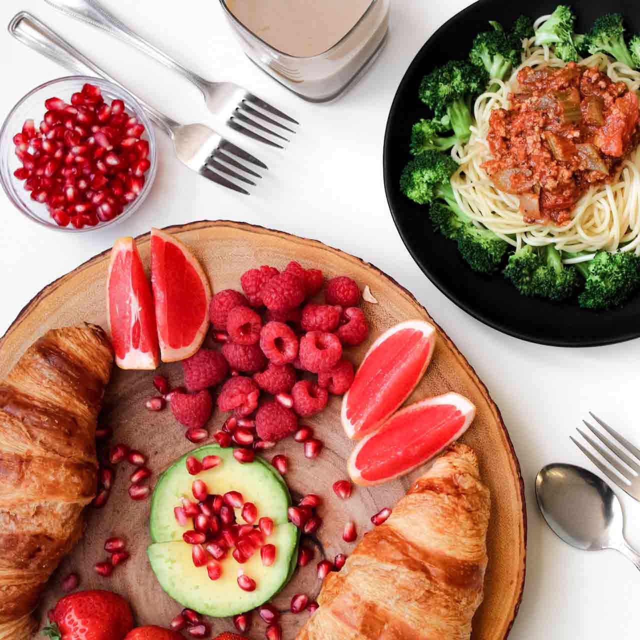 Gedekte tafel met kleurrijk eten. Croissants, fruit, komkommer, pasta met saus en broccoli.